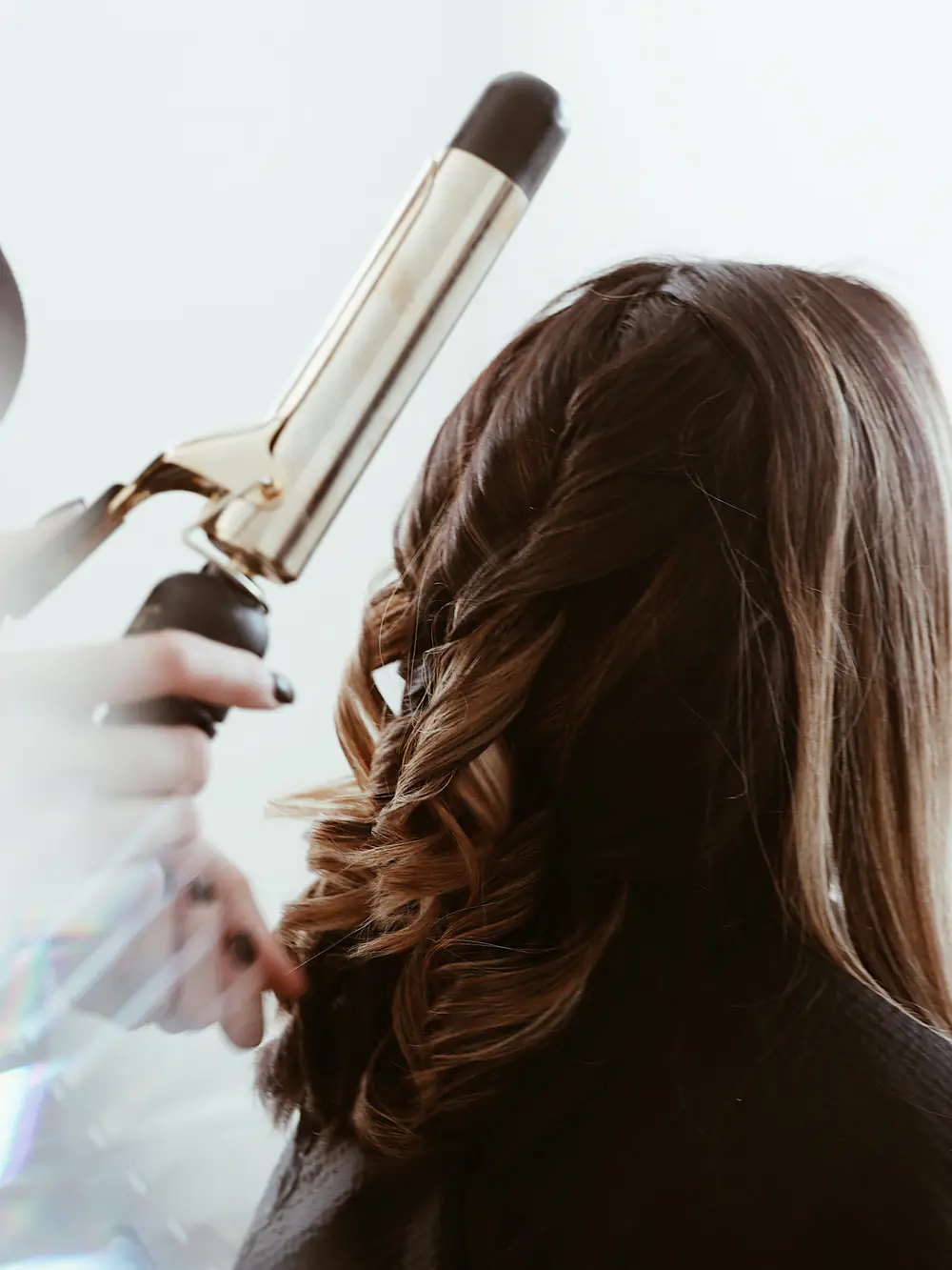 Une coiffeuse boucle au fer une longue chevelure brune, vue de dos