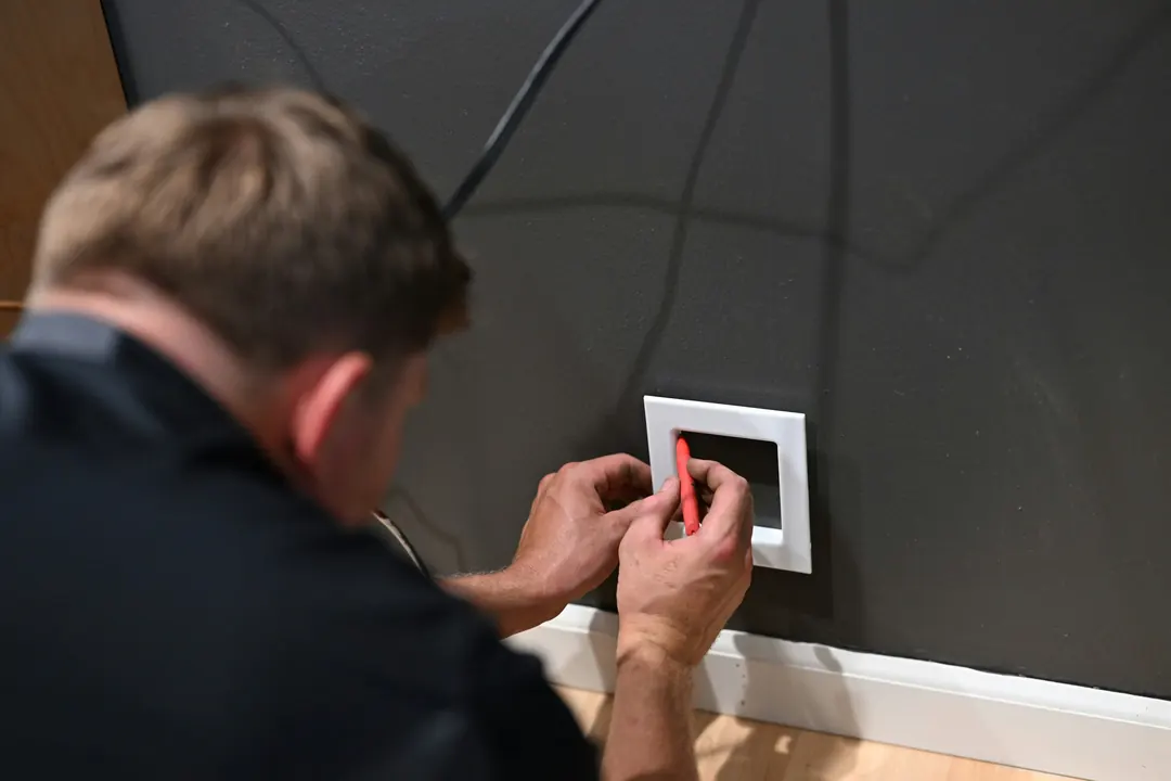Un électricien, vu de dos, ajuste la plaque d’un interrupteur sur un mur gris